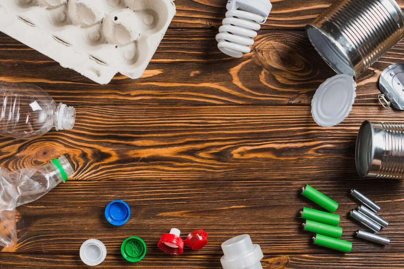 An egg carton, a light bulb, a can, a battery, a cap, and bottles on a wooden textured background.